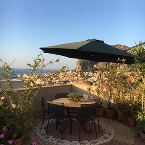 Une terrasse avec une table et des chaises, entourée de plantes en pot, donnant sur une ville côtière avec ses bâtiments et une falaise.