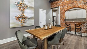 A dining area with a wooden table and grey chairs, a large abstract painting on the wall, and a brick archway leading to a kitchen.