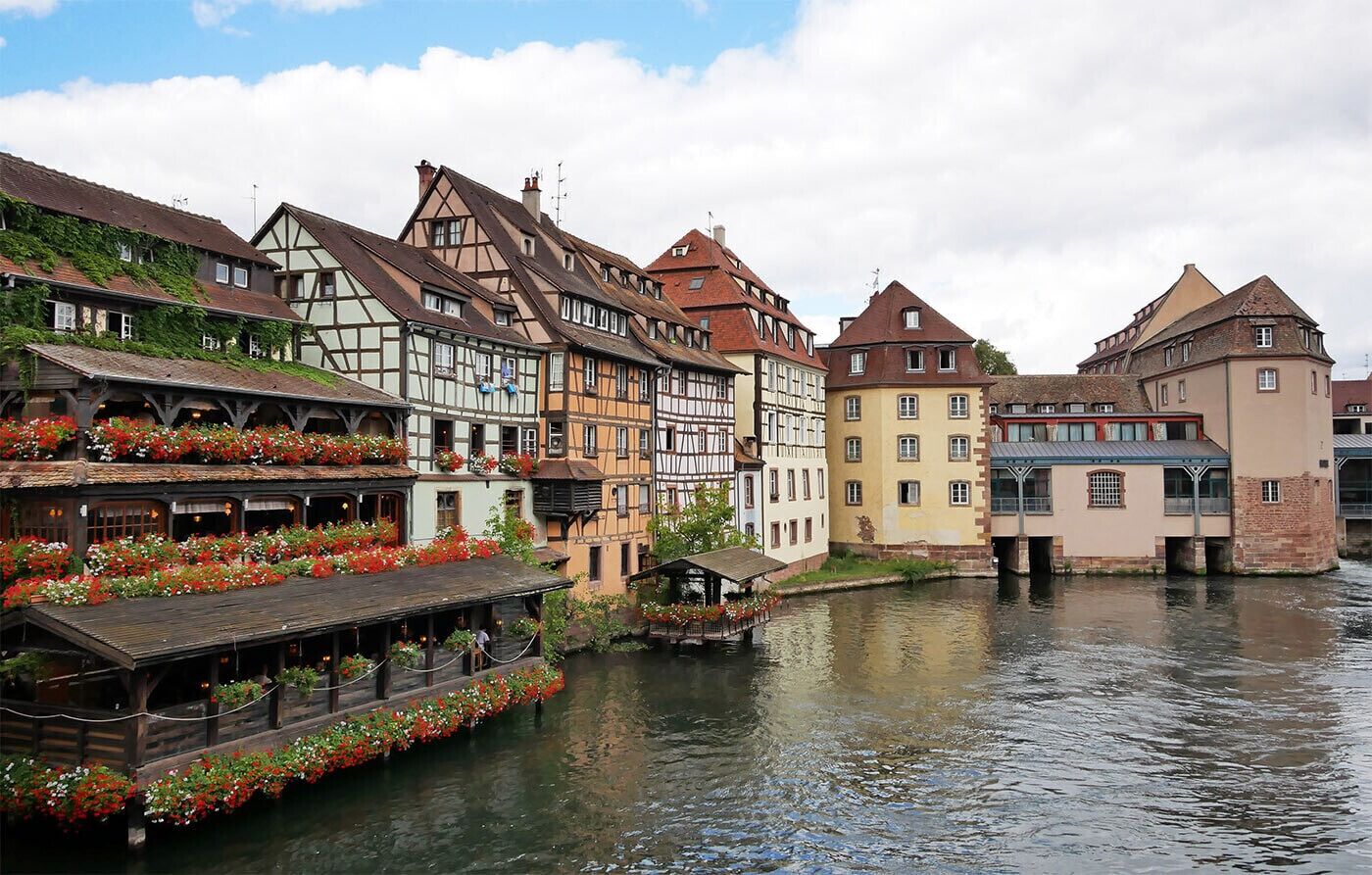 A riverside town with traditional half-timbered buildings, a wooden boat with flower boxes, and a small covered structure.