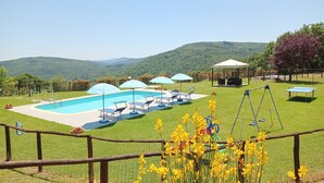 A pool area with lounge chairs and umbrellas, surrounded by a grassy lawn and a view of hills.