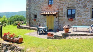 A stone building with a tiled roof, a small porch with a table and chairs, and a pool area.