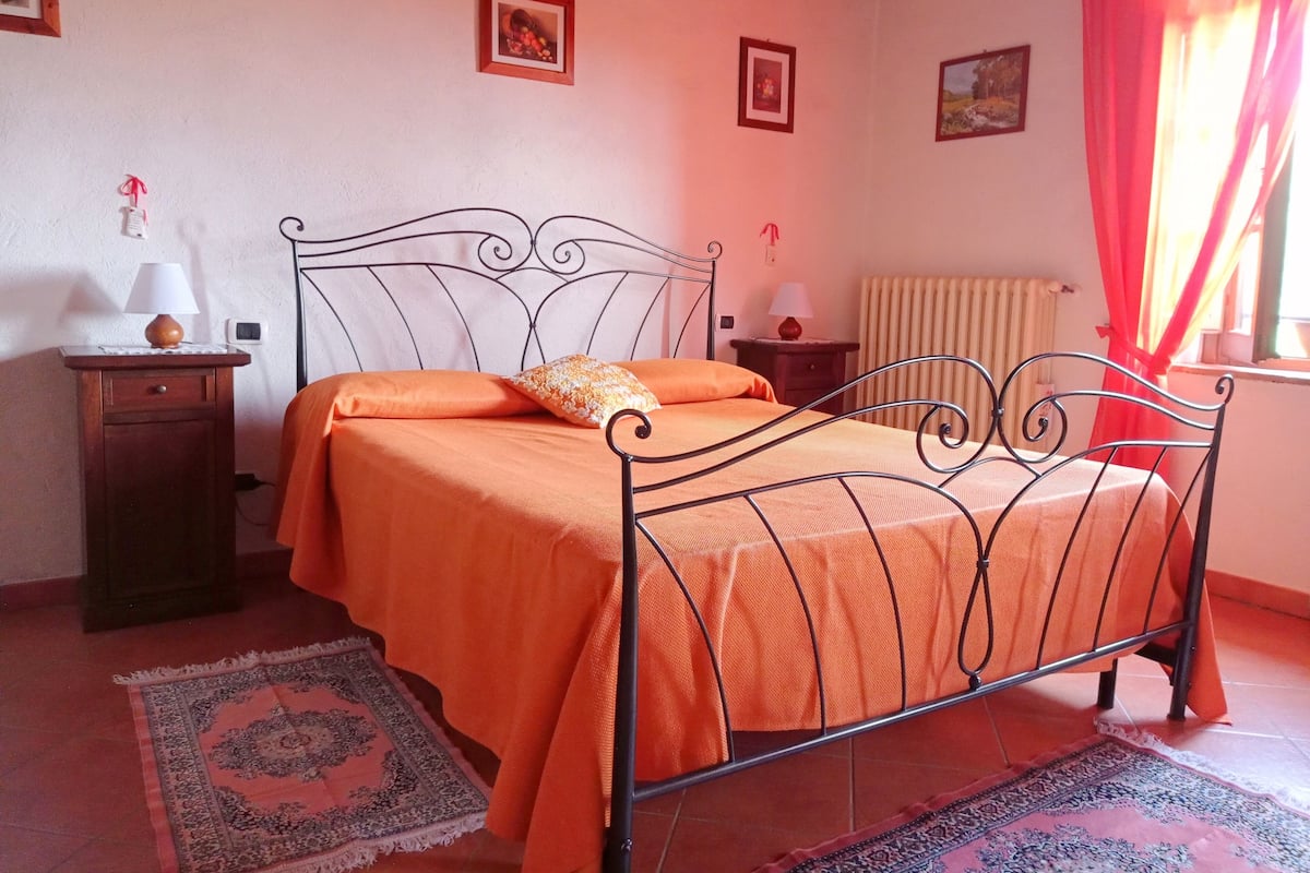 Une chambre avec un lit en métal, une couvre-lit orange, une petite table de chevet avec une lampe et un tapis à motifs sur le sol.