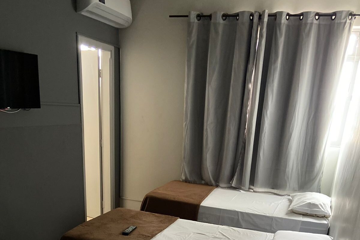A hotel room with two single beds, a wooden headboard, a small table with a remote control, and a window with grey curtains.