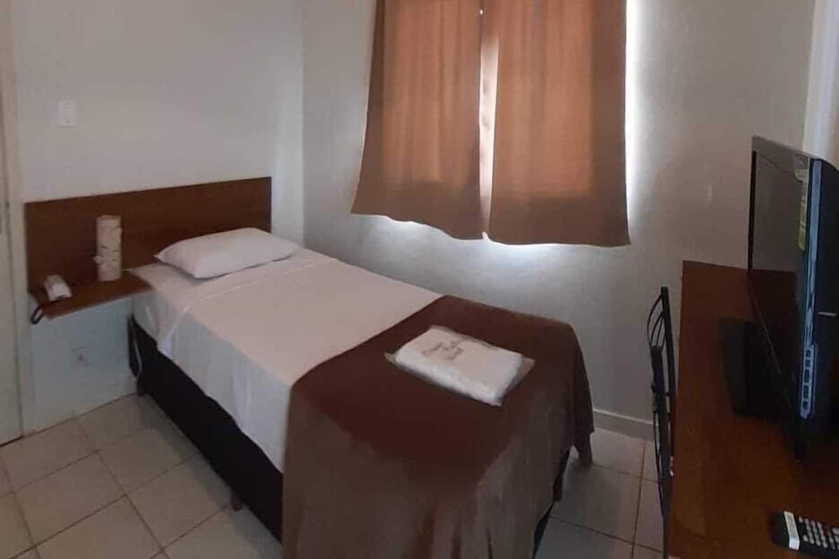 Una cama individual con cabecera de madera, una mesita de noche con lámpara, un televisor y una ventana con cortinas.