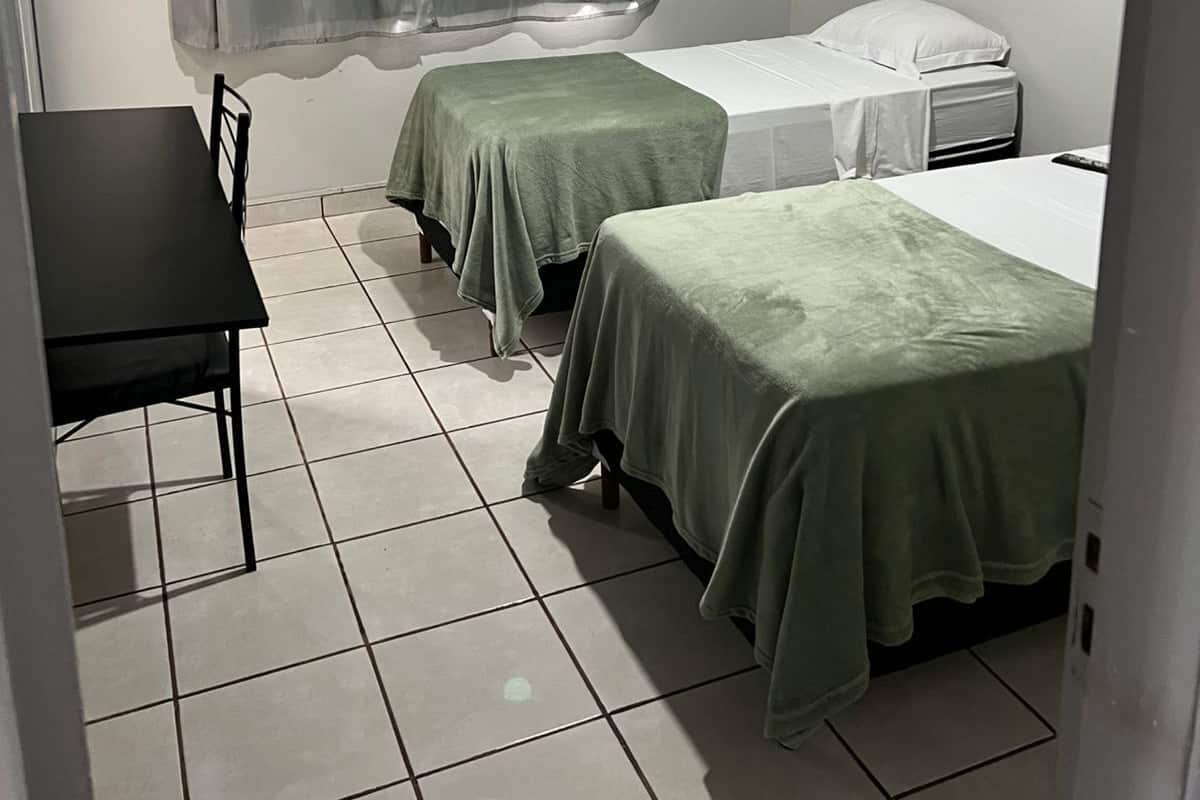 Una habitación pequeña y limpia con dos camas individuales, una mesa y un ventilador de techo.