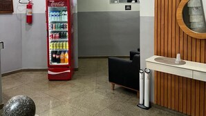 Un área de recepción con una máquina expendedora de Coca-Cola, un extintor, una zona de estar negra, un panel de pared de madera y un espejo redondo.