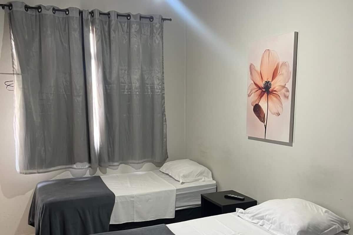 Una habitación con dos camas, un ventilador de techo, una ventana con cortinas y un cuadro de flores colgado en la pared.