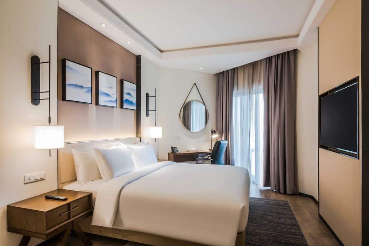 A hotel room with a large bed, bedside tables, a desk, a chair, a TV, and a window with curtains.