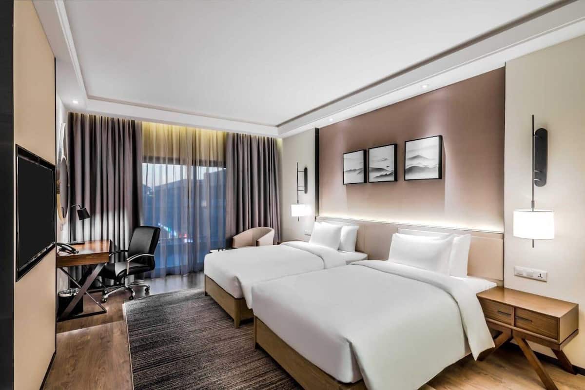 A hotel room with two beds, a desk, a chair, a TV, and a view of the city.