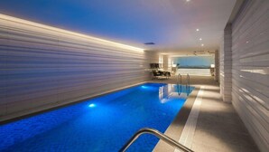 Una piscina interior moderna con un tono azul, rodeada por un diseño minimalista con paredes blancas y un largo pasillo.