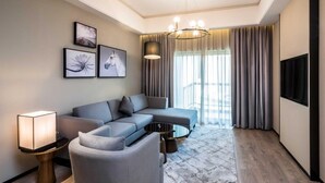 Una habitación de hotel moderna con un sofá gris, una pequeña mesa redonda, una lámpara de pie y un televisor en la pared.
