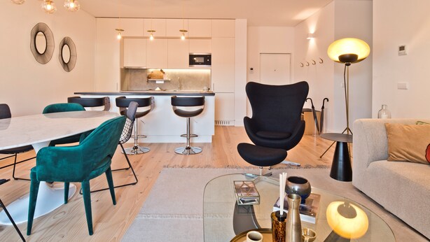 Ein modernes Wohnzimmer mit einem Glastisch, einem schwarzen Sessel und einer Couch. Es gibt einen Essbereich mit Tisch und Stühlen, und im Hintergrund eine Küche.