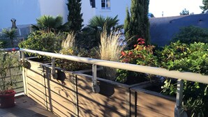 A wooden deck with a railing, potted plants, and a white building in the background. - Charming 2-room terrace ideally located in a quiet area - beautiful unobstructed view. (La Baule)