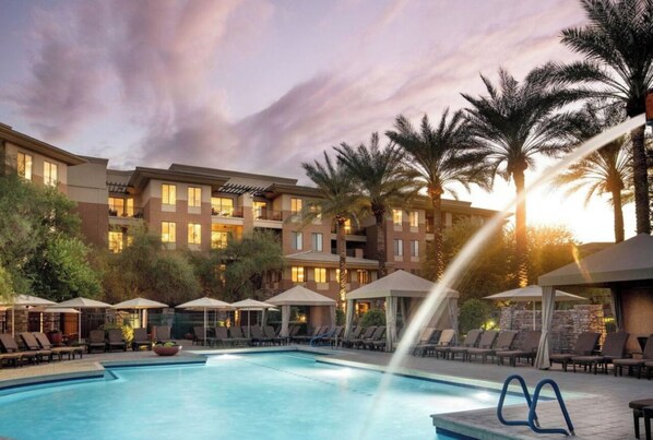 A hotel pool area with sun loungers and umbrellas, surrounded by palm trees and a building with multiple balconies.