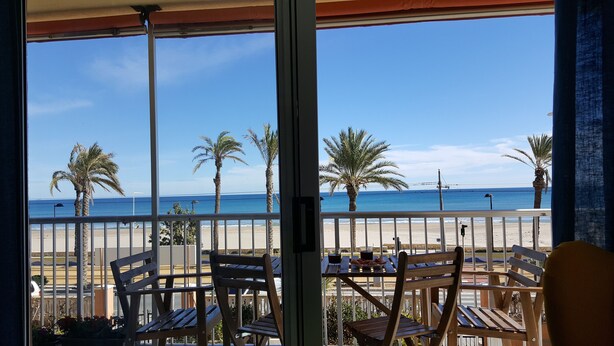 Un balcón con sillas y mesas de madera, con vista a una playa con palmeras y el océano.