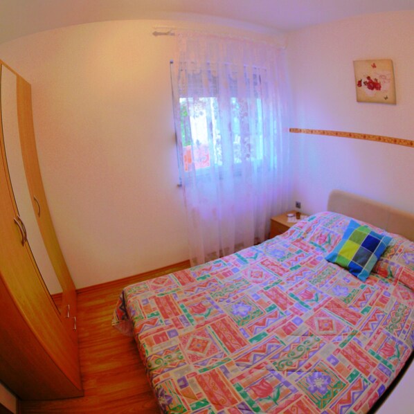 Une chambre à coucher comprenant un lit, une table de chevet, une armoire en bois et une fenêtre avec des rideaux légers.