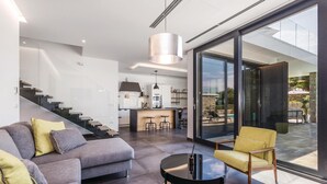 Een moderne woonruimte met een bank, een ronde salontafel en een gele fauteuil. In de achtergrond is een keuken zichtbaar, en er is een glazen deur die leidt naar een buitenruimte.