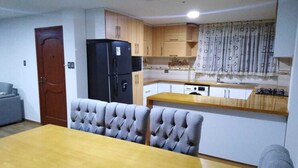 Una cocina compacta con armarios de madera, un refrigerador negro y una zona de comedor con una mesa de madera y sillas tapizadas en gris.