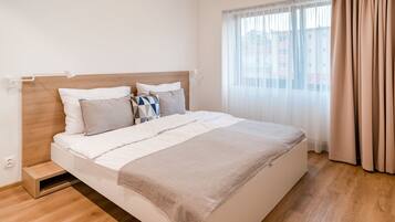 Een moderne hotelkamer met een houten hoofdeinde, een groot bed met witte beddengoed, een beige plaid en een raam met dunne gordijnen.