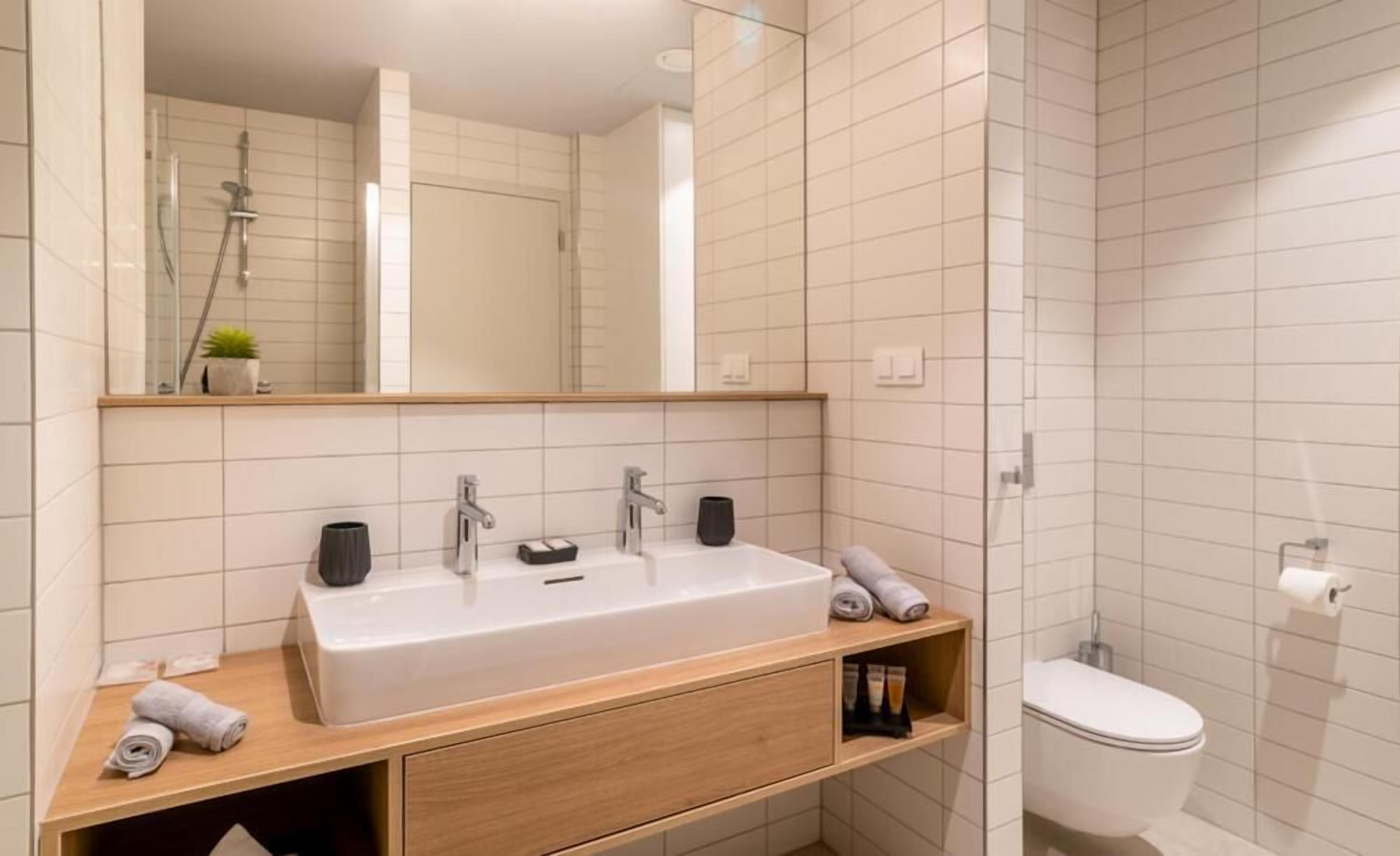 A modern bathroom with a large mirror, a sink with two faucets, a toilet, and a shower area.