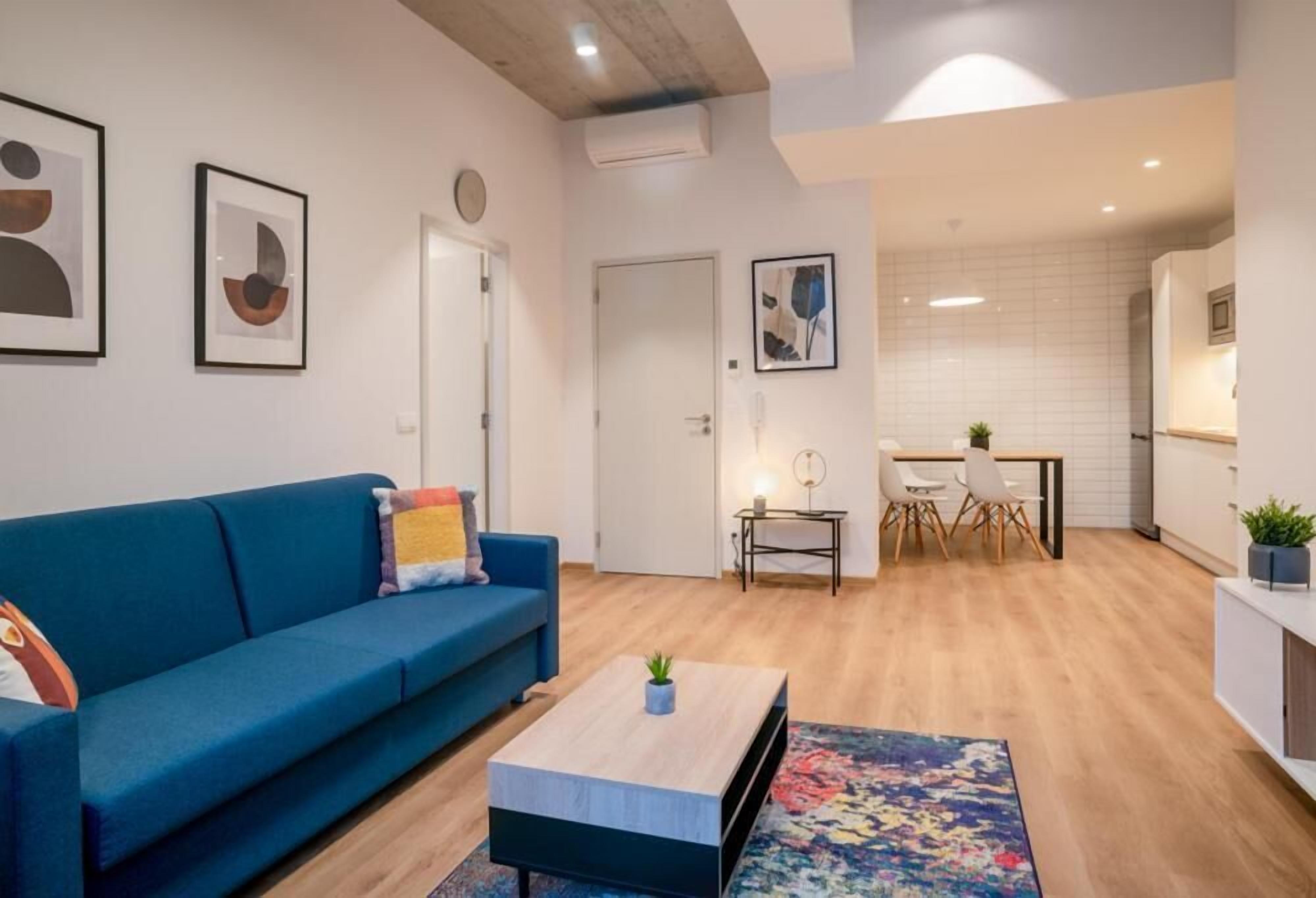 A modern living room with a blue sofa, a coffee table, and a colorful rug. There is a dining area with a white table and chairs, and a kitchen in the background.