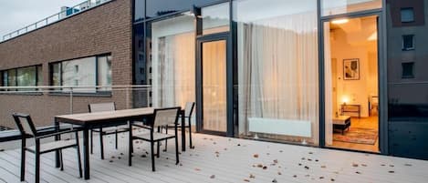 A modern building with a wooden deck, a dining area with chairs, and large glass doors leading to an interior room.