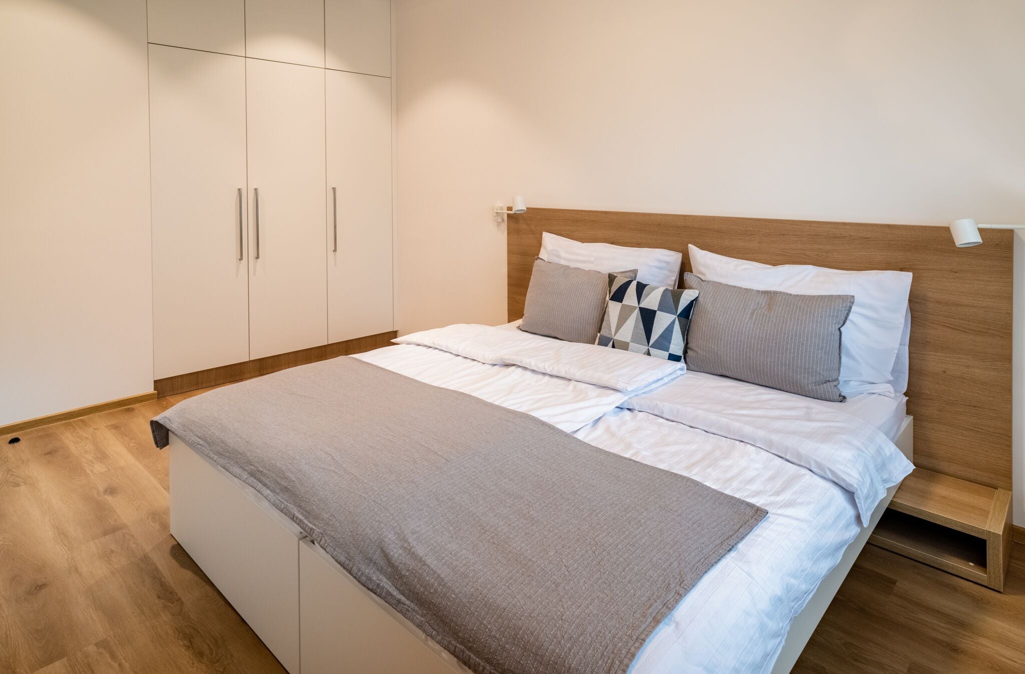 A neatly made bed with a wooden headboard, a grey blanket, and multiple pillows. There is a white wardrobe with silver handles and a wooden floor.