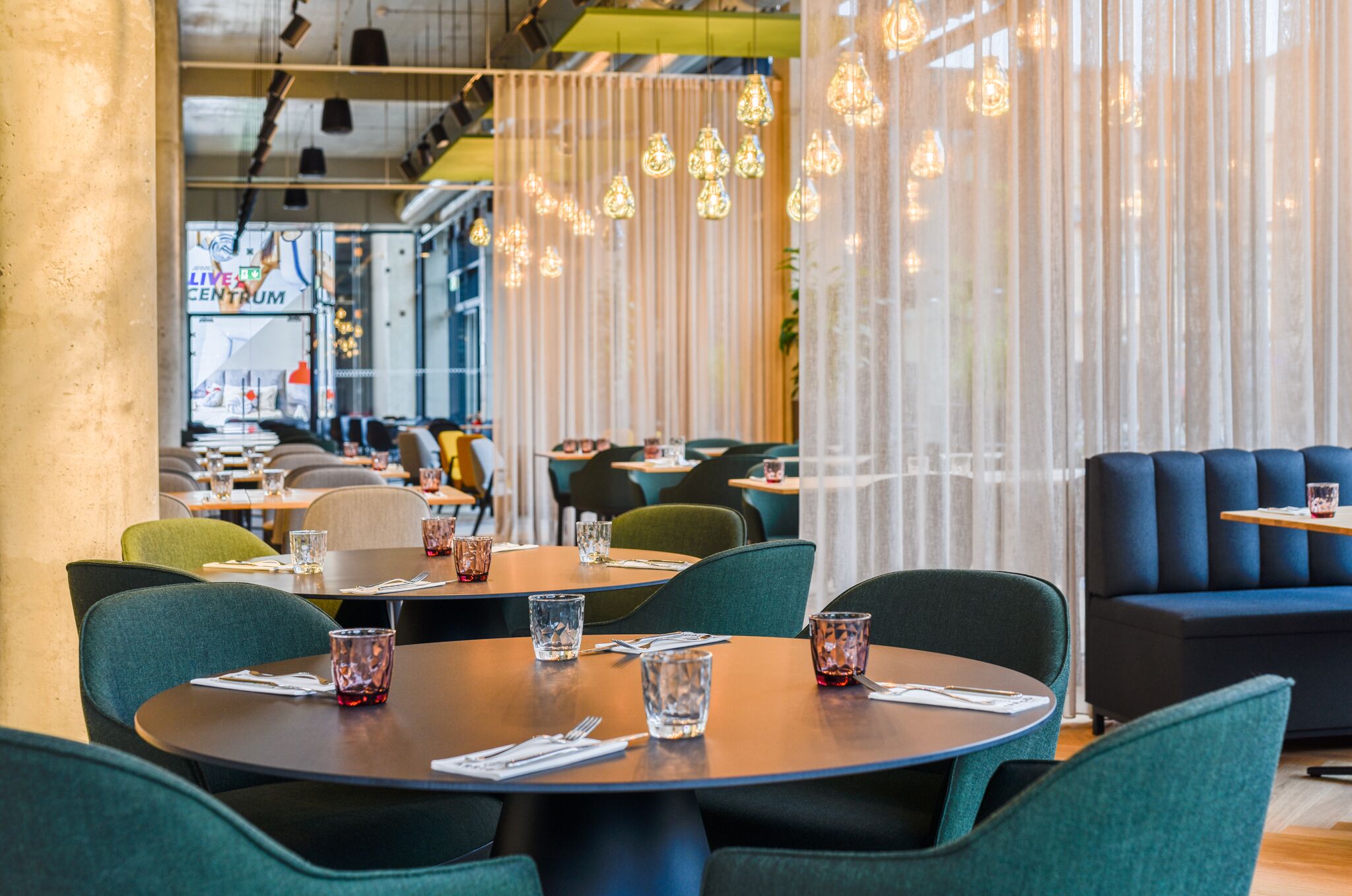 A modern restaurant interior with round tables, green upholstered chairs, and hanging pendant lights.