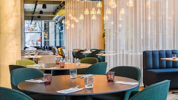 A modern restaurant interior with round tables, green upholstered chairs, and hanging pendant lights.