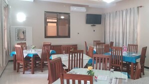 A dining area with tables and chairs, a television on the wall, and a window with blinds. - Kigala Hotel (Dar es Salaam)