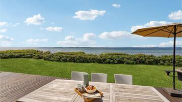 Une terrasse en bois avec une table et des chaises, une table dressée avec un saladier de fruits et un journal, et une vue sur la mer.
