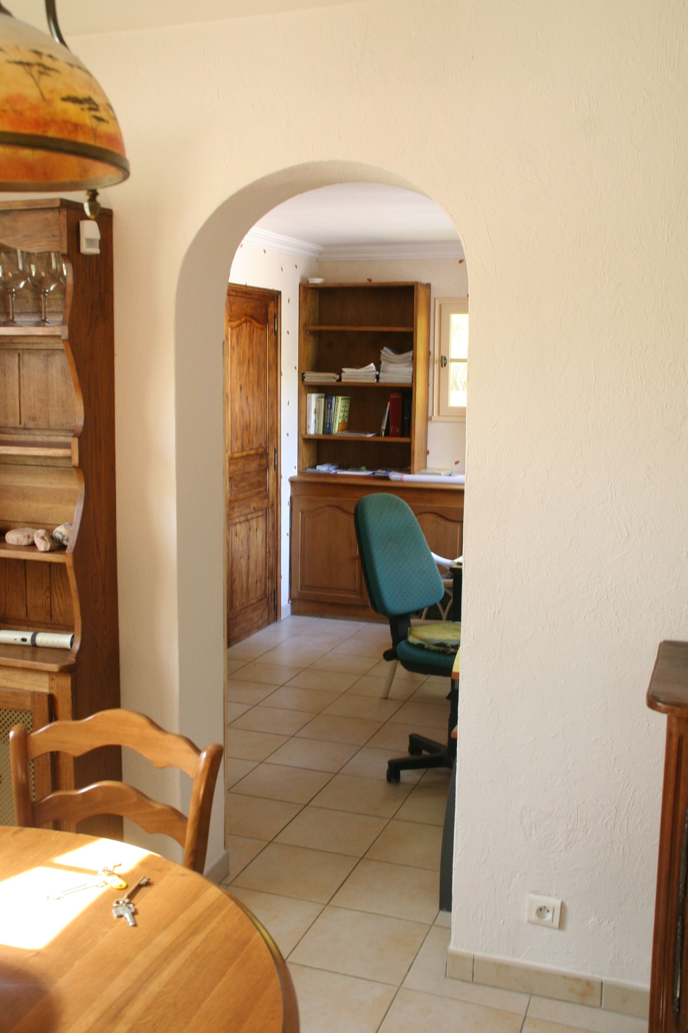 Un comedor con una mesa de madera, sillas y una lámpara colgante. Se ve una cocina al fondo, con armarios de madera y una silla.