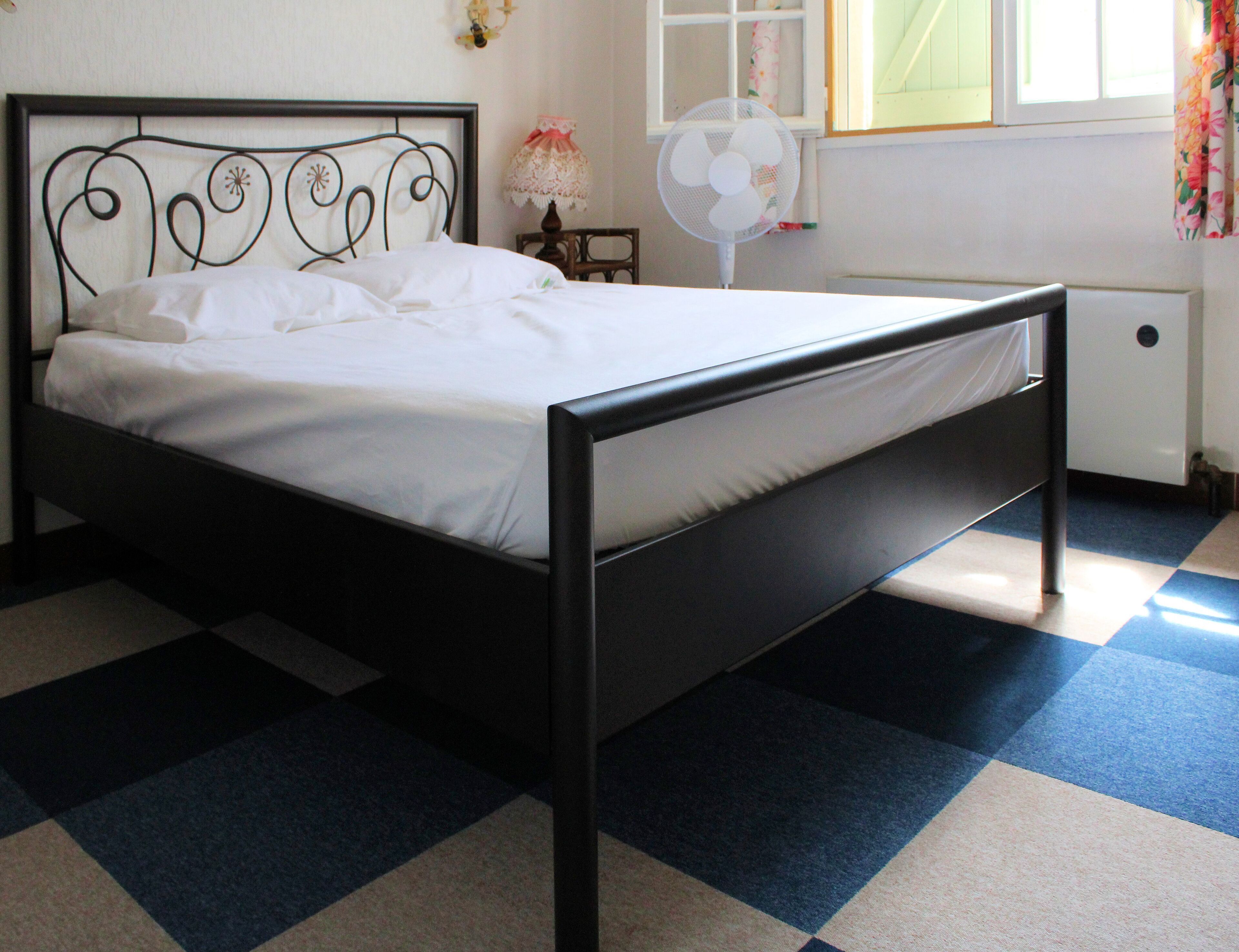 A bed with a metal frame and white bedding, a standing fan, and a window with floral curtains.