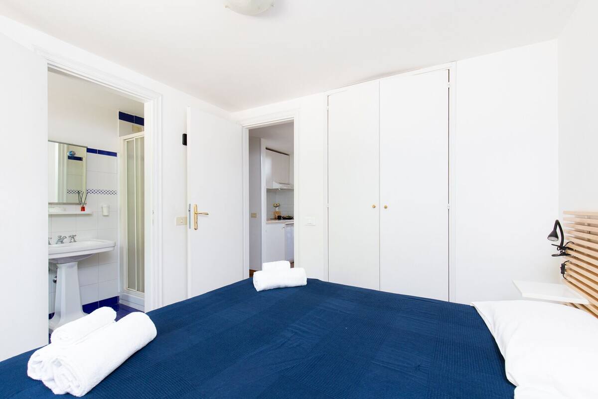 A bedroom with a bed, a bedside table, a mirror, a wardrobe, and a bathroom with a sink and mirror.