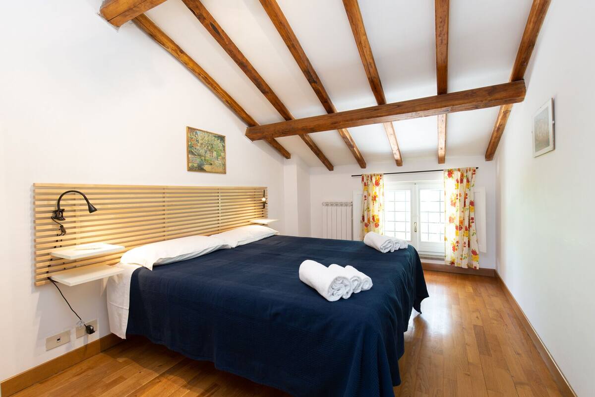 A bedroom with a wooden ceiling, a bed with a dark blue bedspread, a bedside table with a lamp, and a window with floral curtains.