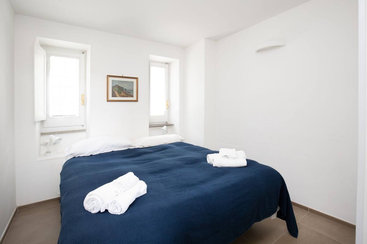 A hotel room with a large bed, two windows, a painting on the wall, and a towel on the bed.