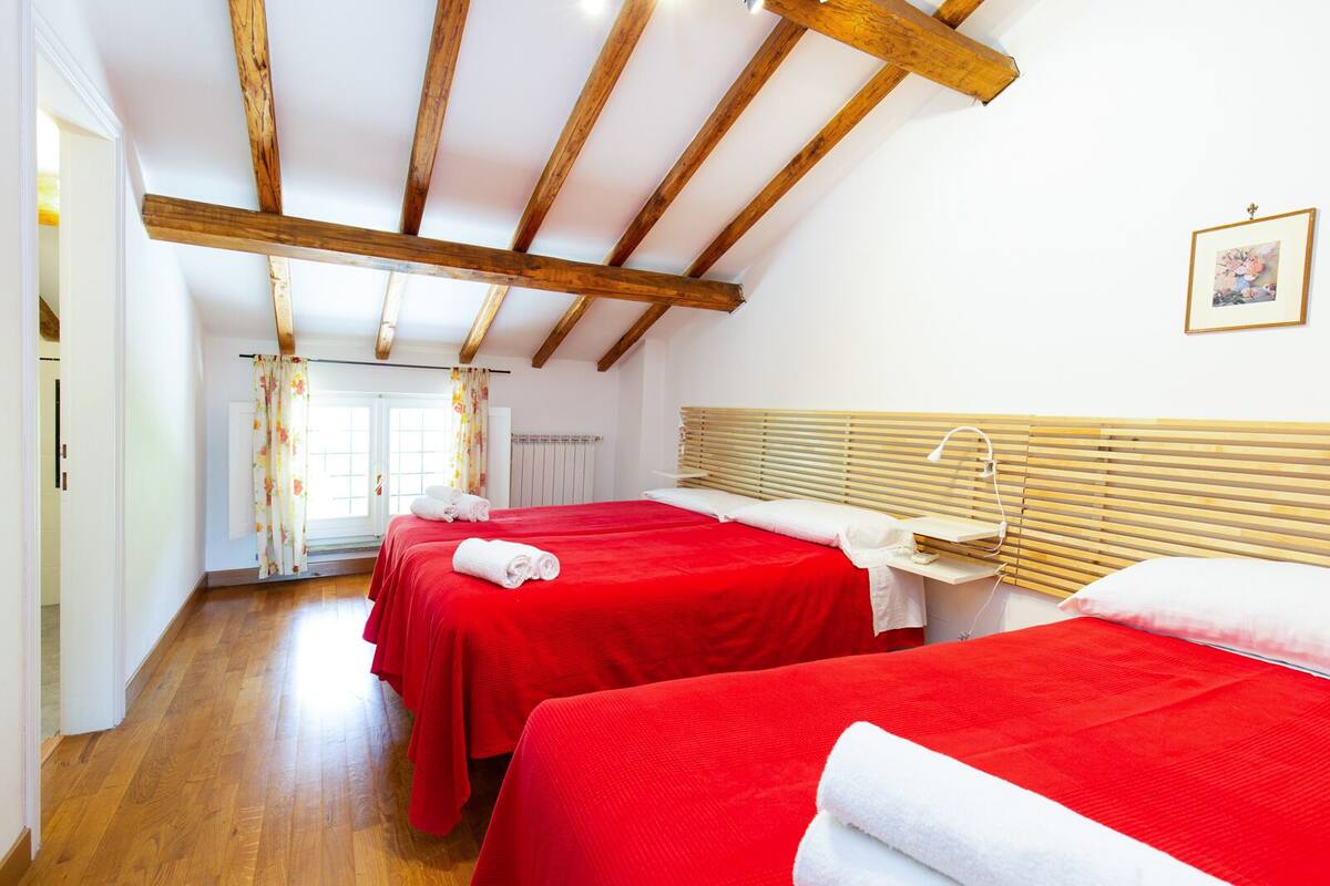 A room with two beds, wooden beams on the ceiling, a window with curtains, and a radiator.