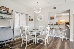 Zona pranzo moderna con tavolo in vetro e sedie bianche, con una cucina sullo sfondo e un lampadario a sospensione.