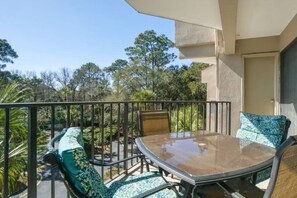 Un balcon doté d’une table et de chaises à plateau en verre, entouré d’une rambarde en métal, et offrant une vue sur un jardin aménagé avec des arbres et des arbustes.