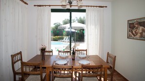 Un coin repas avec une table dressée pour quatre personnes, des chaises en bois, un lustre et une vue sur une piscine et des parasols à l’extérieur.