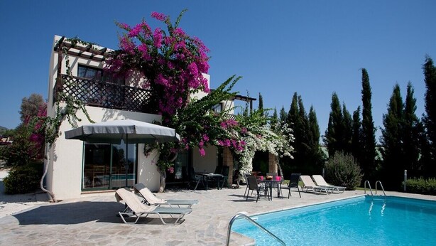 Un espace aménagé au bord d’une piscine à l’eau d’un bleu limpide, avec des chaises longues et un bâtiment doté d’un balcon orné de fleurs roses.