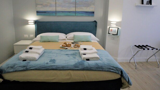 A neatly made bed with a blue headboard, a bedside table with a lamp, and a wall-mounted shelf with a picture. - Porta San Gennaro (Naples)