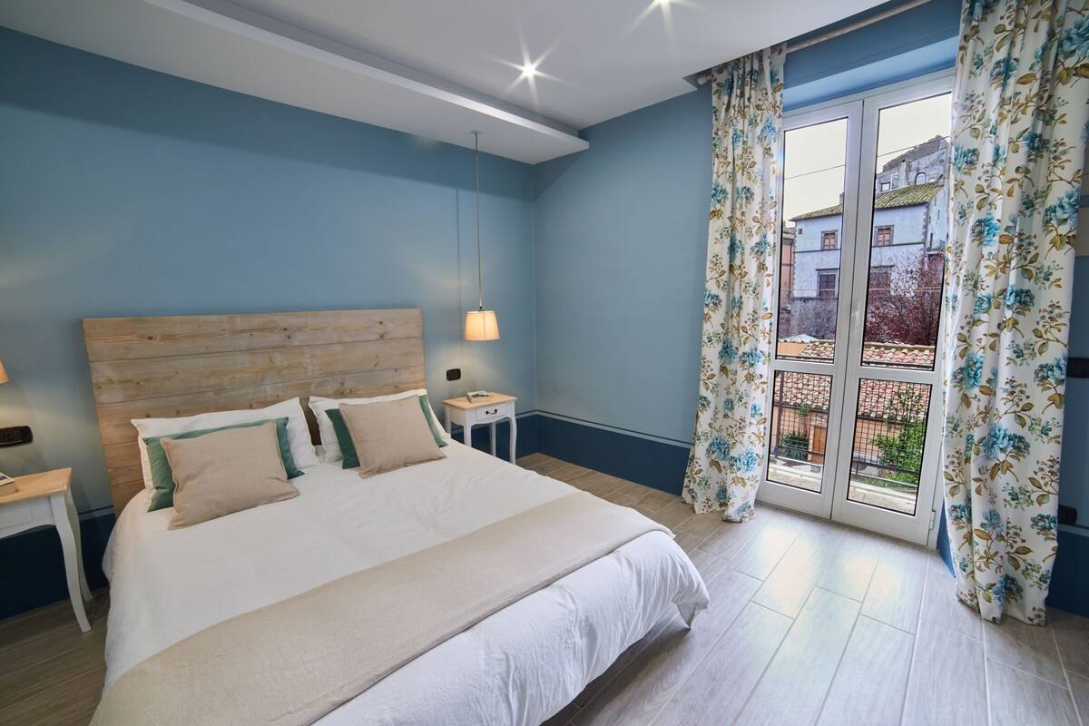 A bedroom with a wooden headboard, a large bed with white bedding, a nightstand, a lamp, and a window with floral curtains.