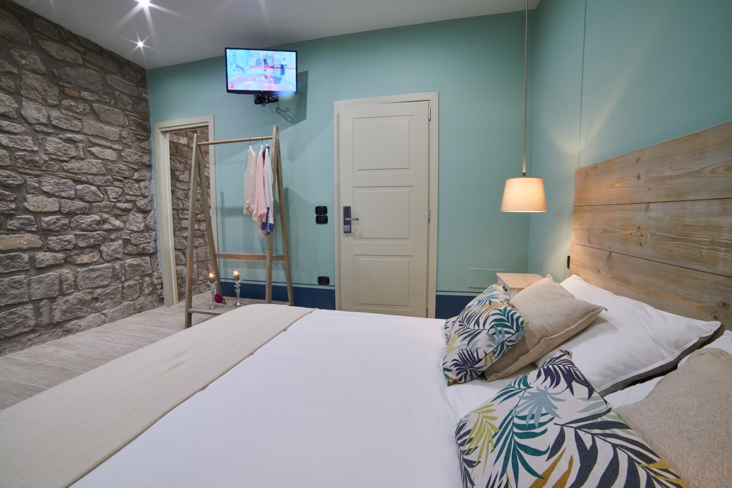 Un dormitorio con pared de piedra, cabecera de madera, cama con almohadones estampados, televisor en la pared y una escalera apoyada contra la pared.