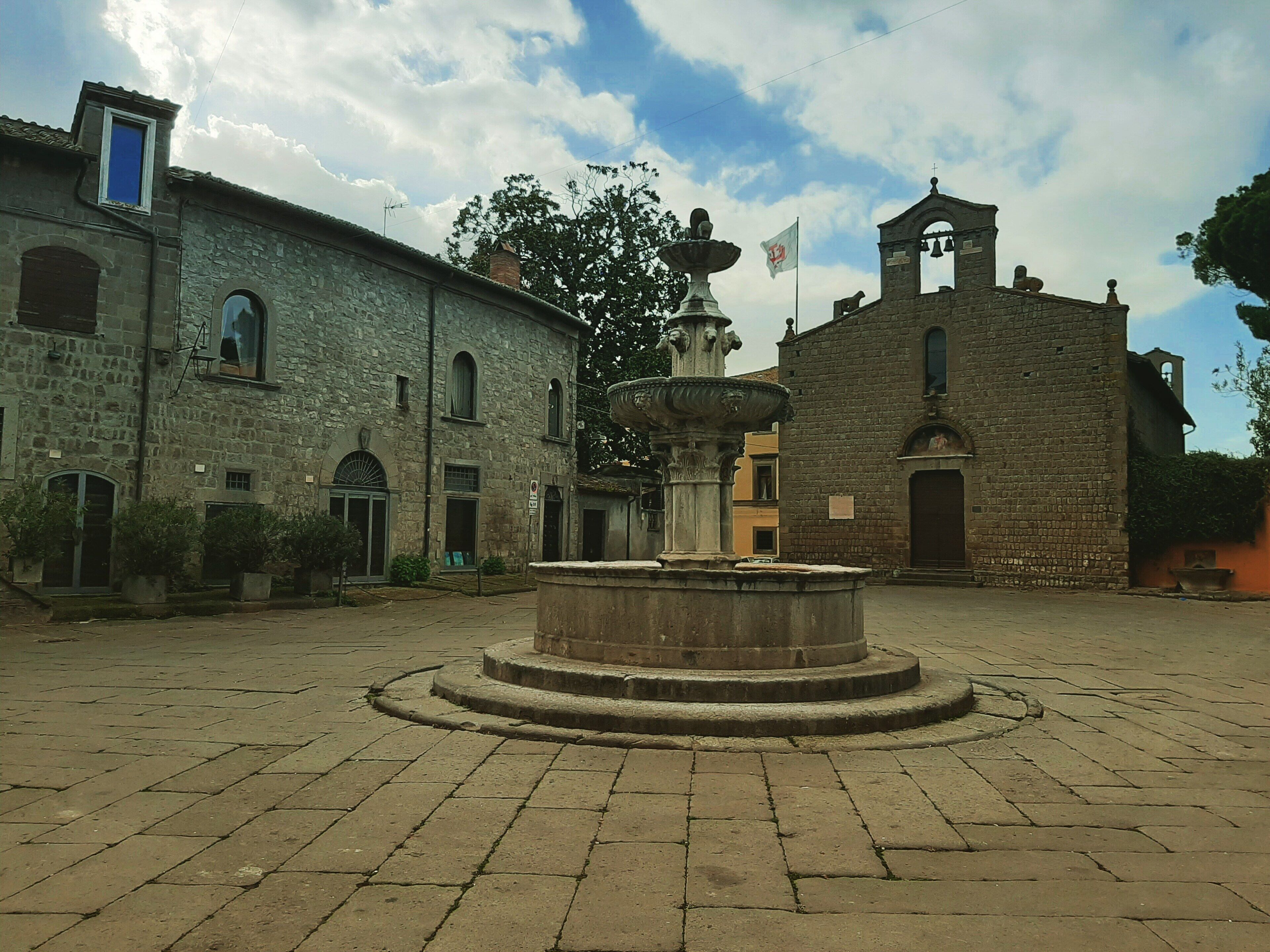 Un cortile con una fontana al centro, edifici in pietra e un campanile.