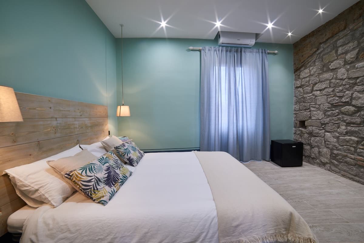 A bedroom with a wooden headboard, a bed with white bedding, a patterned pillow, a wall-mounted lamp, a stone wall, and a window with curtains.