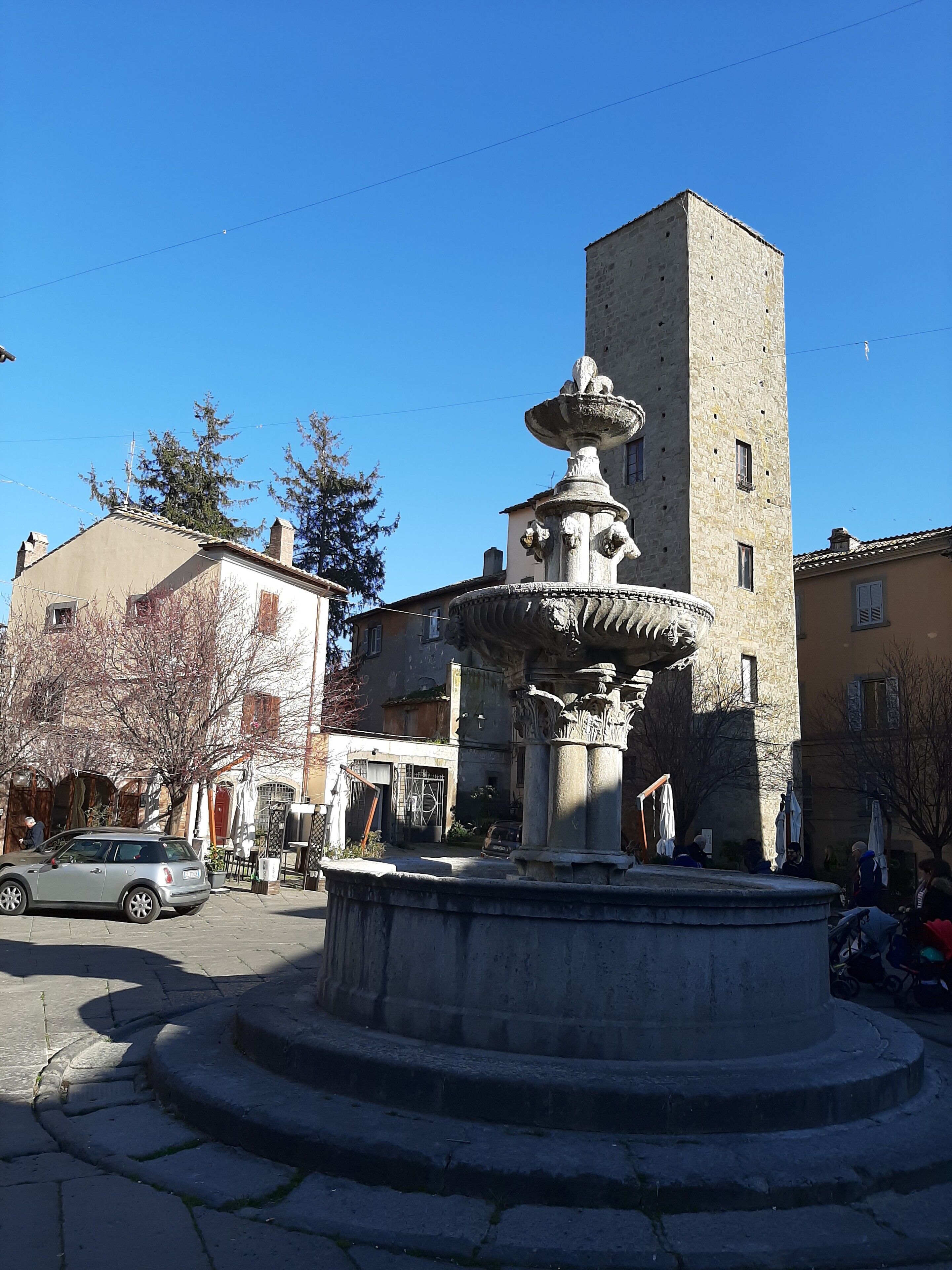 A stone fountain with a tiered design in a square, surrounded by buildings with traditional architecture.