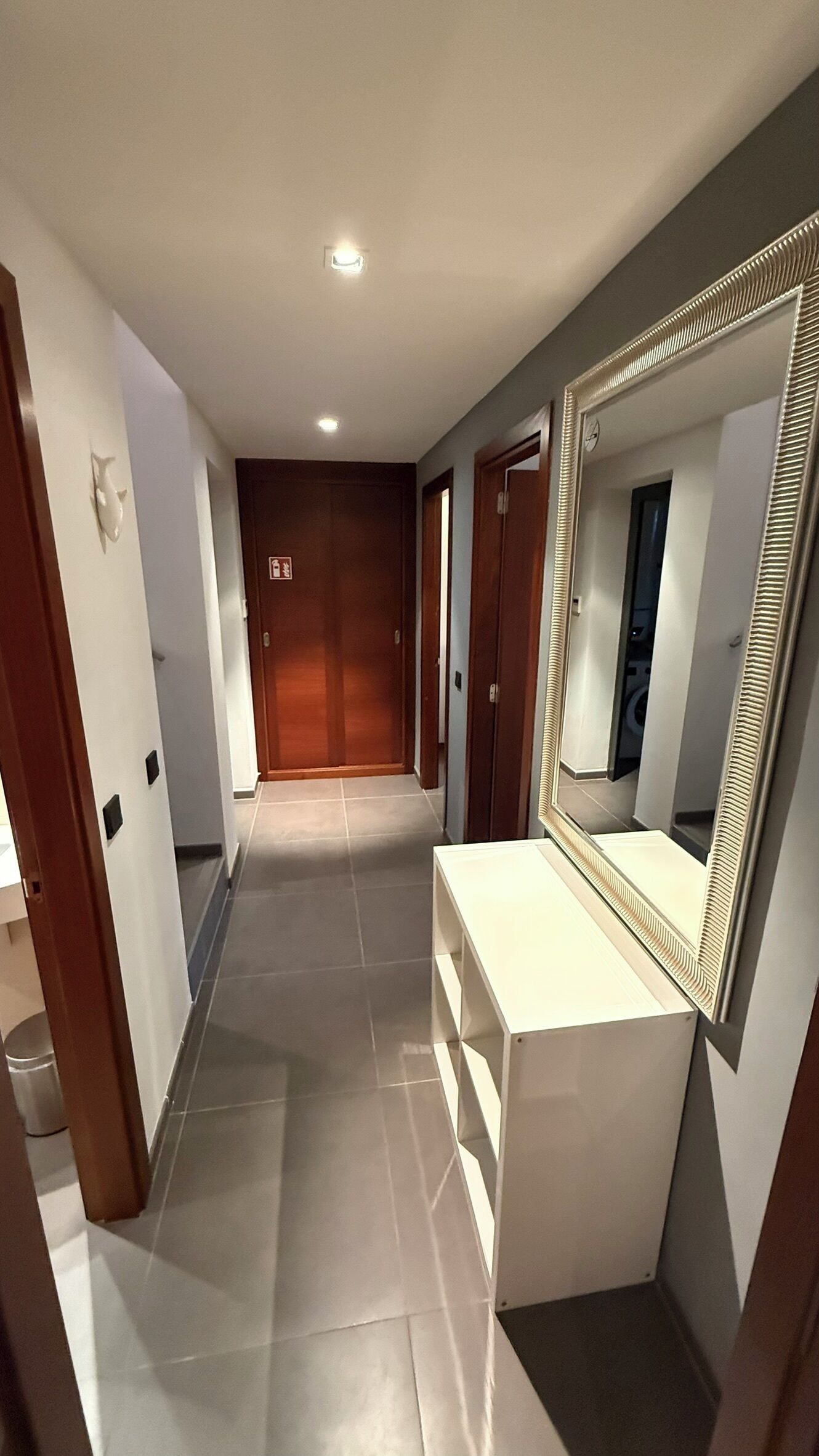 Un couloir avec une coiffeuse blanche, un grand miroir et une porte en bois au fond.