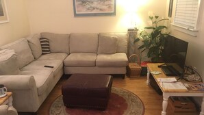 Una sala de estar con un sofá beige, un reposapiés marrón, una mesa de centro de madera, un televisor y una planta.