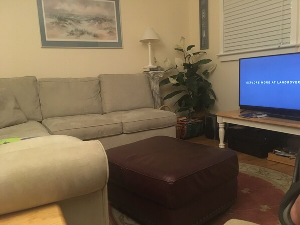 Un salon avec un canapé beige, un pouf marron, un téléviseur affichant le message « DÉCOUVREZ-EN PLUS SUR LANDROVER », une plante en pot et une lampe.